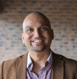 Smiling man in brown suit jacket with red brick wall in background