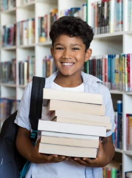 Smiling young boy holding stack of books in library